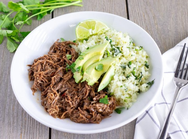 White bowl with shredded mexican beef, cilantro lime cauliflower rice, and topped with sliced avocado.