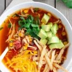 Overhead view of chicken tortilla soup in a white bowl with half an avocado and cilantro leaves on the side.