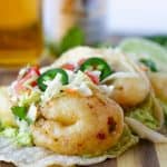 Beer Battered shrimp tacos topped with slaw and jalapenos on a wood board with a glass of Modelo on the side.