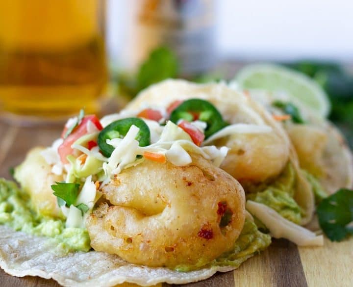 Beer Battered shrimp tacos topped with slaw and jalapenos on a wood board with a glass of Modelo on the side.