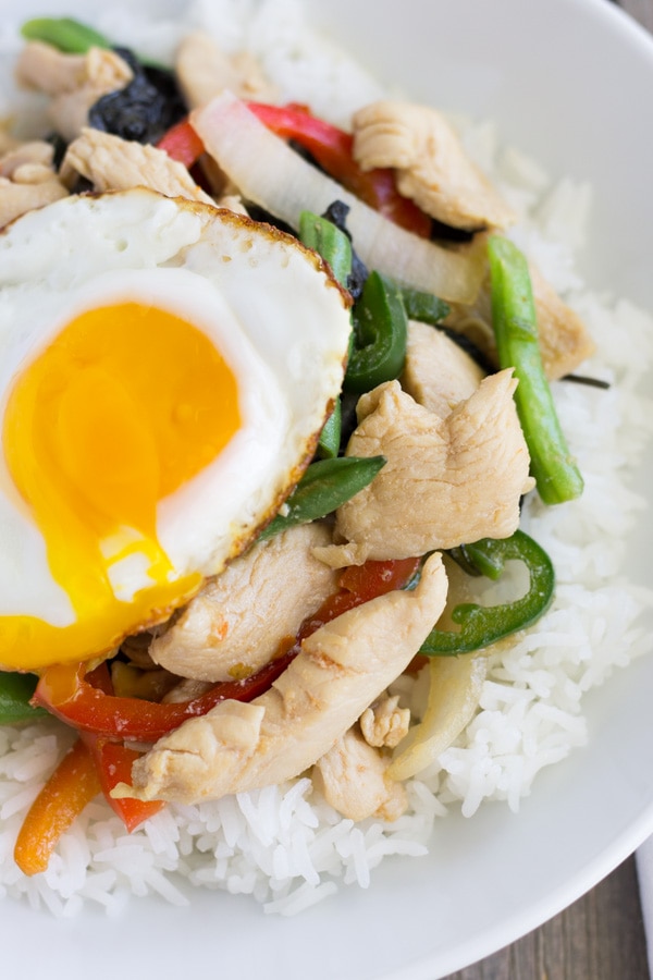 White bowl with rice on the bottom and spicy basil chicken on top with a fried egg and runny yolk.