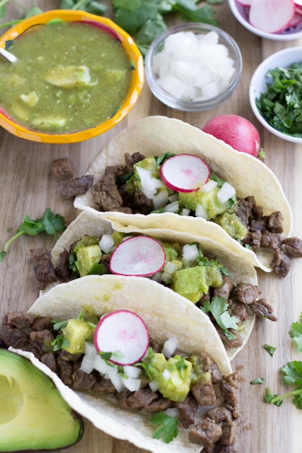 Three tacos on a cutting board topped with salsa verde, cilantro, onions, and radishes.
