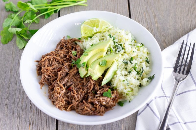 White bowl with shredded mexican beef, cilantro lime cauliflower rice, and topped with sliced avocado.